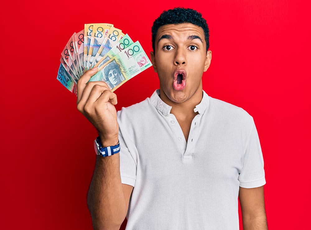Young man holding australian dollars scared and amazed with open mouth for surprise, disbelief face